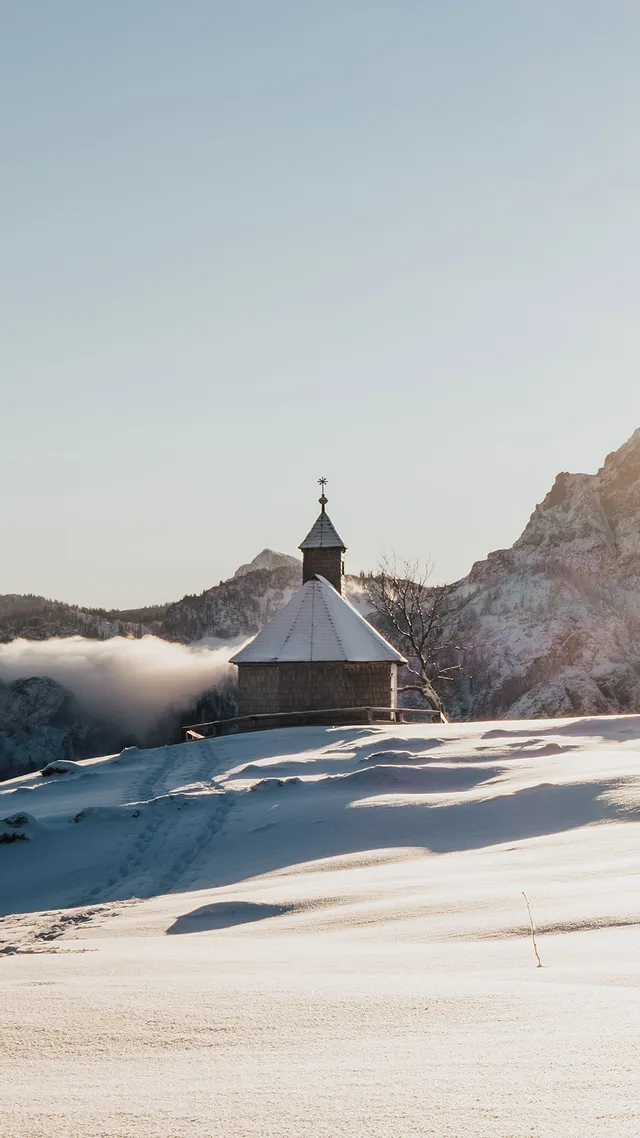 Zwei Wanderer wandern durch verschneite Alpenlandschaft in Richtung Sonnenaufgang und Bergkapelle