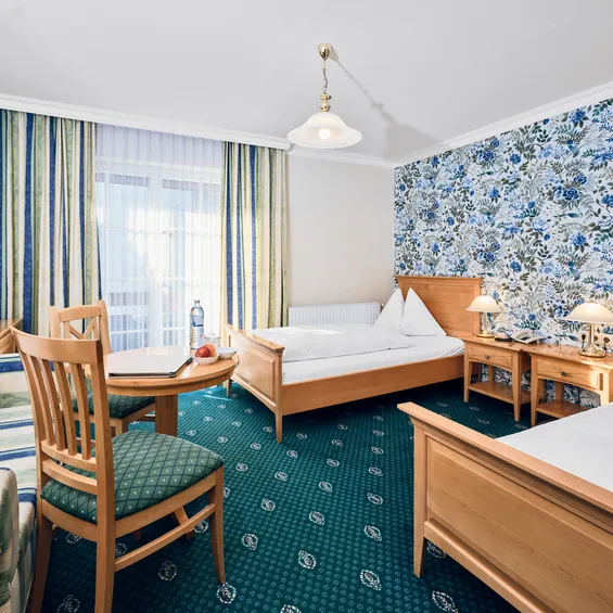 Bright hotel room with two single beds, floral wallpaper, table, chairs, and a TV