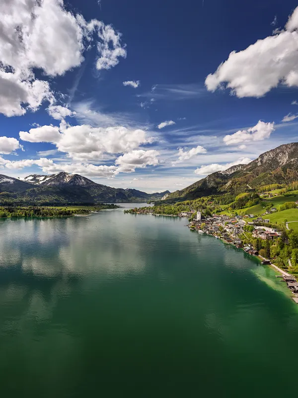 Malerisches Dorf am See mit grünen Hügeln und schneebedeckten Bergen unter teils bewölktem Himmel