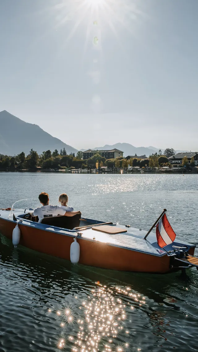 Couple on wooden motorboat cruising on alpine lake with mountain and village view