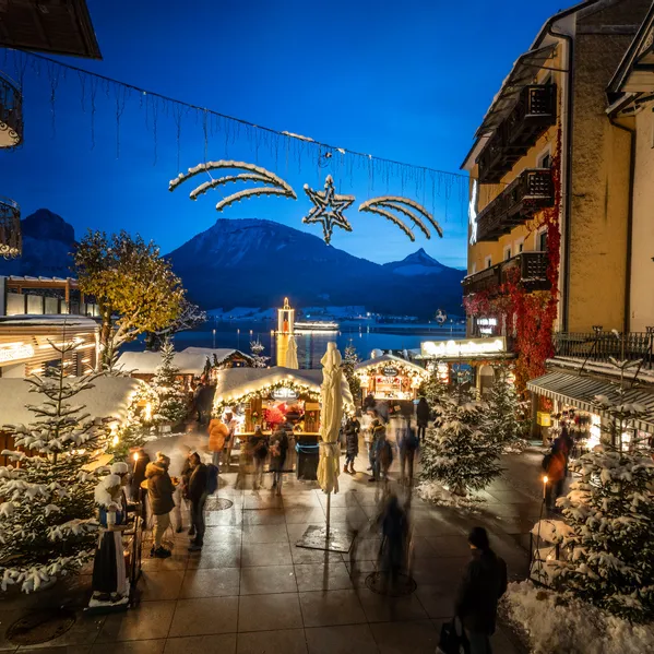 Festlicher Weihnachtsmarkt mit Lichtern und Schnee auf einem Stadtplatz bei Dämmerung in den Bergen