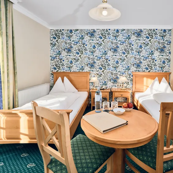 Helles Hotelzimmer mit zwei Einzelbetten, Blumenmuster-Tapete und Holztisch mit Wasser und Obst