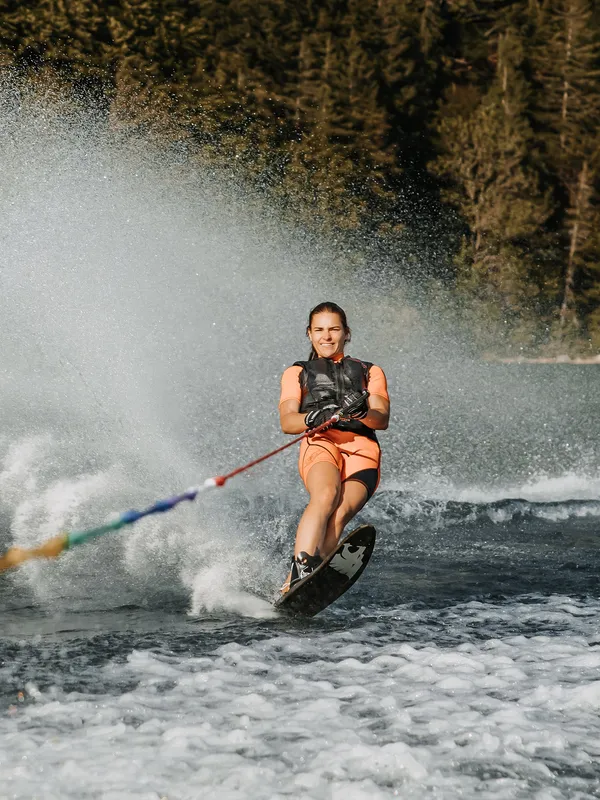 Frau wakeboardet auf einem See mit Wald im Hintergrund und Wasserspritzern rundherum