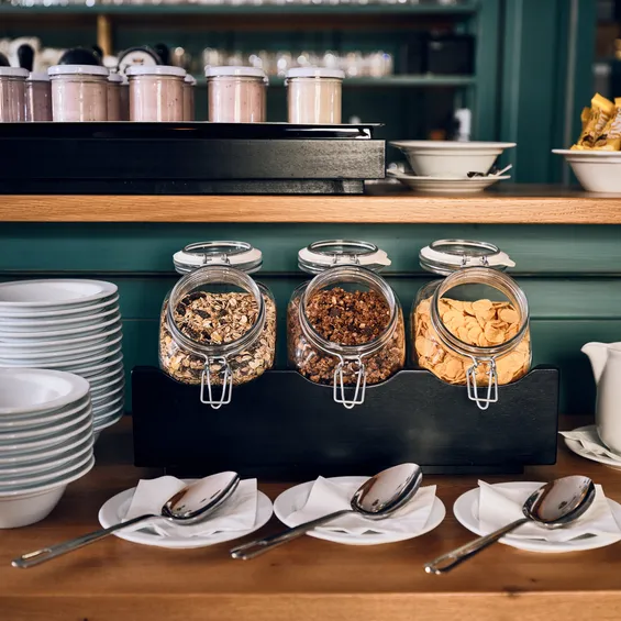 Frühstücksbuffet mit Müsligläsern, gestapelten Schüsseln, Löffeln, Milchkanne und Müsliriegeln