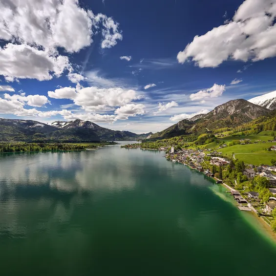 Scenic lakeside village with green hills and snow-capped mountains under a partly cloudy sky