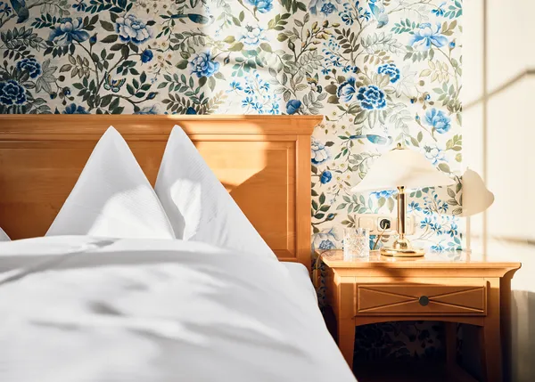 Gemütliches Hotelbett mit weißer Bettwäsche und Blumenmuster-Tapete im Morgenlicht