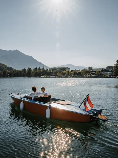 Paar auf Holzmotorboot fährt über Alpensee mit Blick auf Berge und Dorf