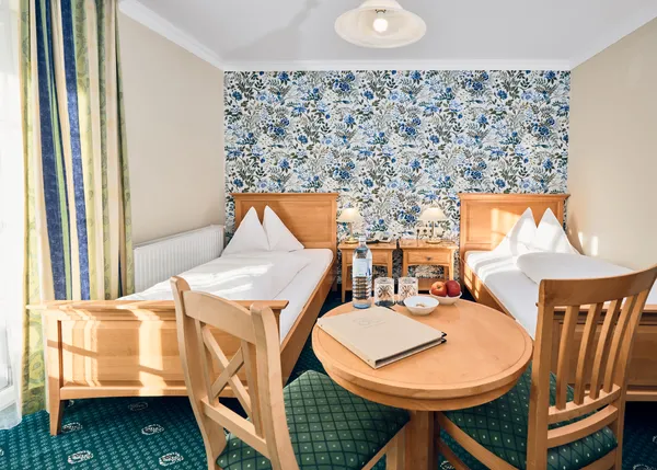 Helles Hotelzimmer mit zwei Einzelbetten, Blumenmuster-Tapete und Holztisch mit Wasser und Obst