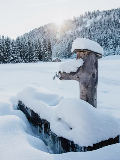 Verschneiter Holzbrunnen in alpiner Winterlandschaft mit Tannen und Bergen