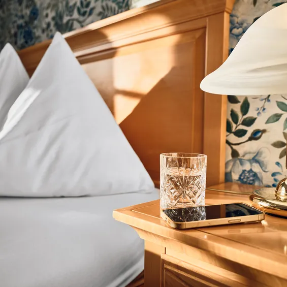 Gemütliches Hotelbett mit weißen Kissen und Nachttisch mit Smartphone, Wasserglas und Messinglampe