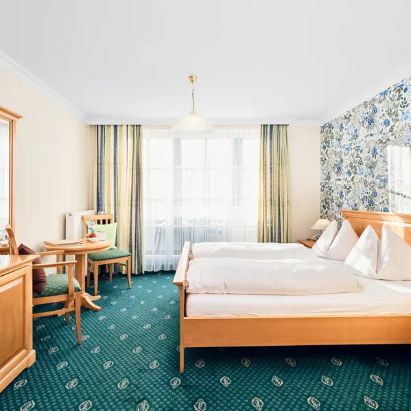 Helles Hotelzimmer mit Doppelbett, Blumenmustertapete, Holzmöbeln und grünem Teppichboden
