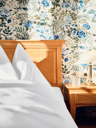 Gemütliches Hotelbett mit weißer Bettwäsche und Blumenmuster-Tapete im Morgenlicht