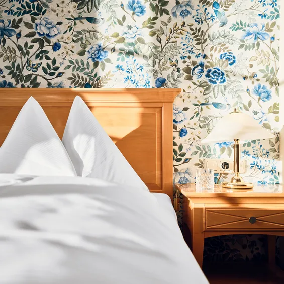 Gemütliches Hotelbett mit weißer Bettwäsche und Blumenmuster-Tapete im Morgenlicht