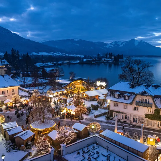 Snow-covered Christmas market by a lake with festive lights and mountain backdrop