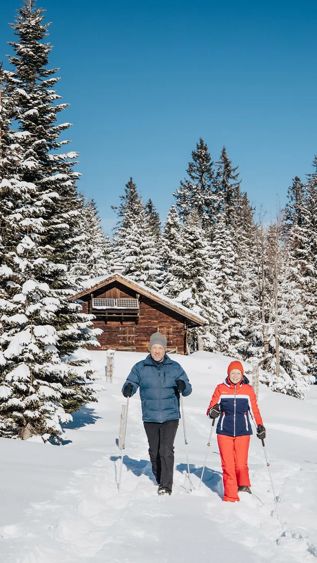 Two people snowshoeing through snowy alpine forest with a wooden cabin in the background
