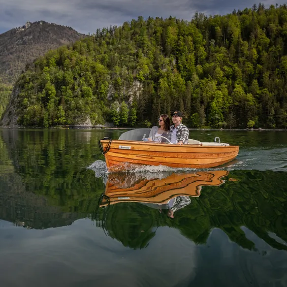 Paar fährt in Holzmotorboot auf ruhigem Bergsee umgeben von Wald